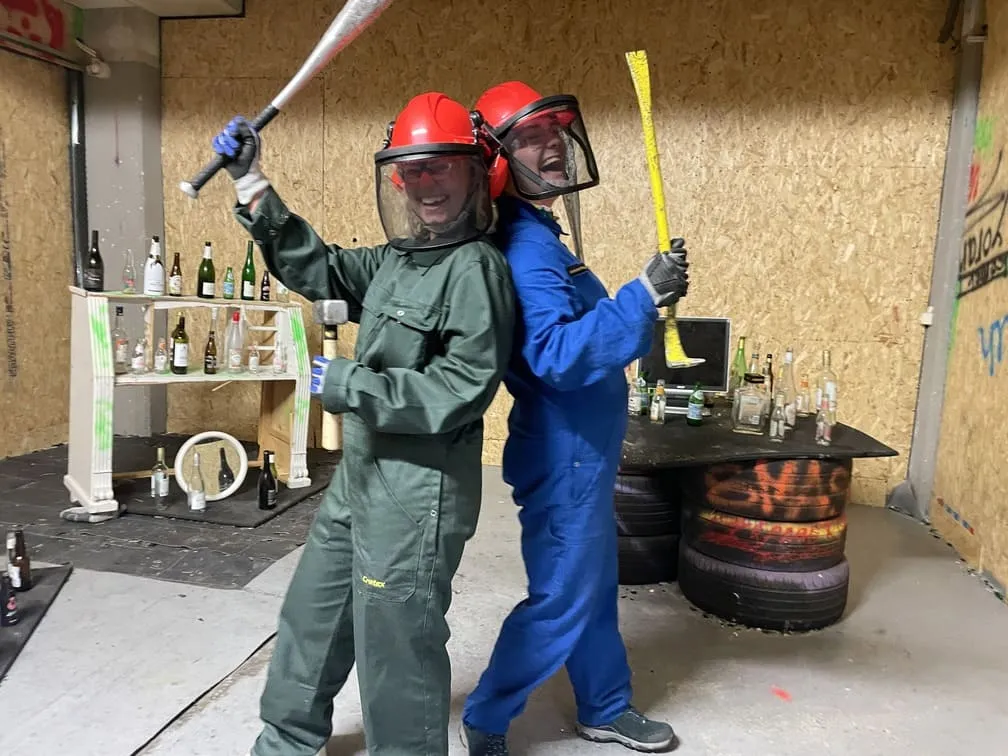 Twee mensen in veiligheidsuitrusting poseren met een knuppel en breekijzer in een ragekamer